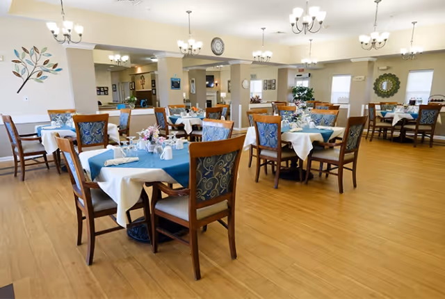 A spacious dining room in a senior living facility with multiple tables covered in white tablecloths and blue runners. Each table is set with napkins, cups, and small floral centerpieces. The room has wooden flooring, chandeliers hanging from the ceiling, and decorative wall art including a tree design and a circular mirror. Large windows allow natural light to brighten the space.