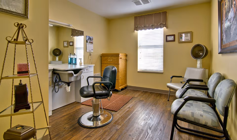 A small hair salon room with a black salon chair in front of a white sink and mirror. There are three waiting chairs with patterned upholstery along the right wall, a vintage hair dryer, a wooden cabinet, and a window with blinds and a valance. The walls are painted yellow and the floor is wood.