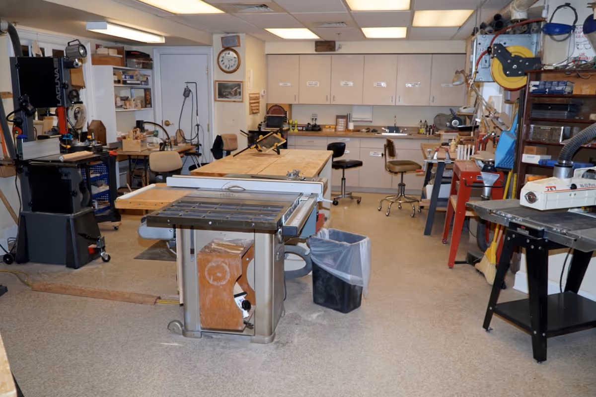 Interior view of a woodworking workshop with various tools and equipment including a table saw, band saw, workbenches, stools, cabinets, and shelves filled with materials and supplies.