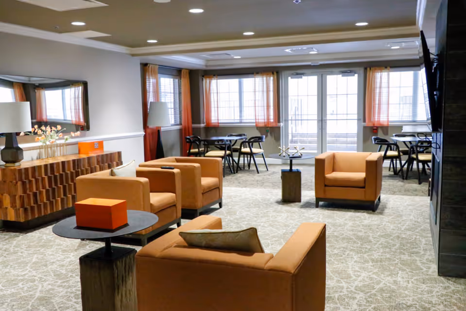 A spacious assisted living common area with beige carpet and neutral walls. The room features four orange armchairs arranged around small tables, a wooden sideboard with a lamp and flowers, and several small dining tables with black chairs near large windows with orange curtains. A large flat-screen TV is mounted on the right wall.