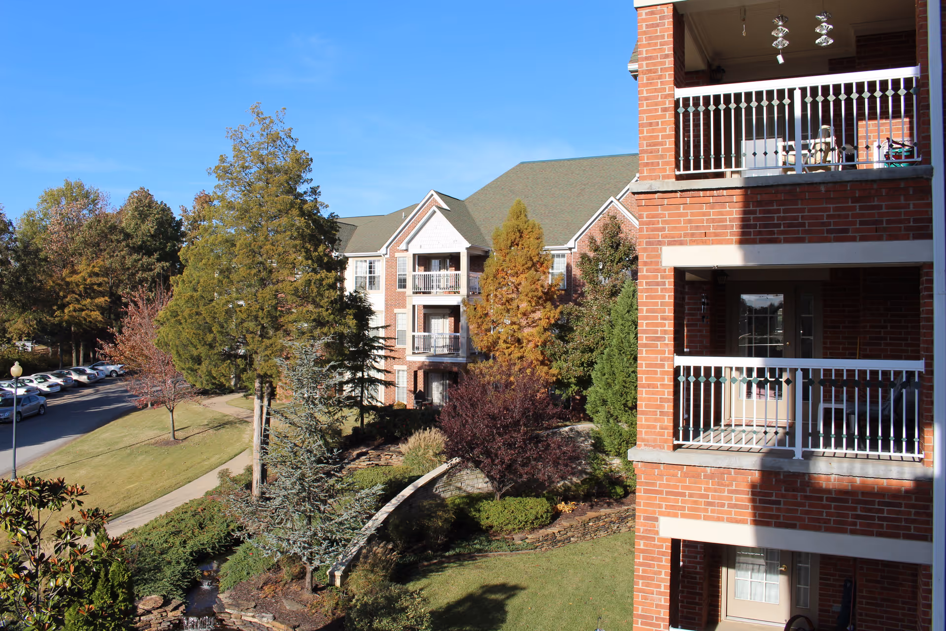 View of a senior living facility named St. Bernards Village showing a multi-story brick building with balconies, surrounded by trees, landscaped gardens, and a parking area with cars under a clear blue sky.
