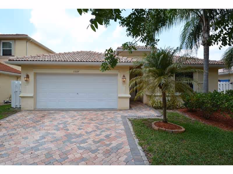 Front exterior view of a single-story house with a tiled roof, a double garage door, a paved driveway, and landscaping including palm trees and bushes.