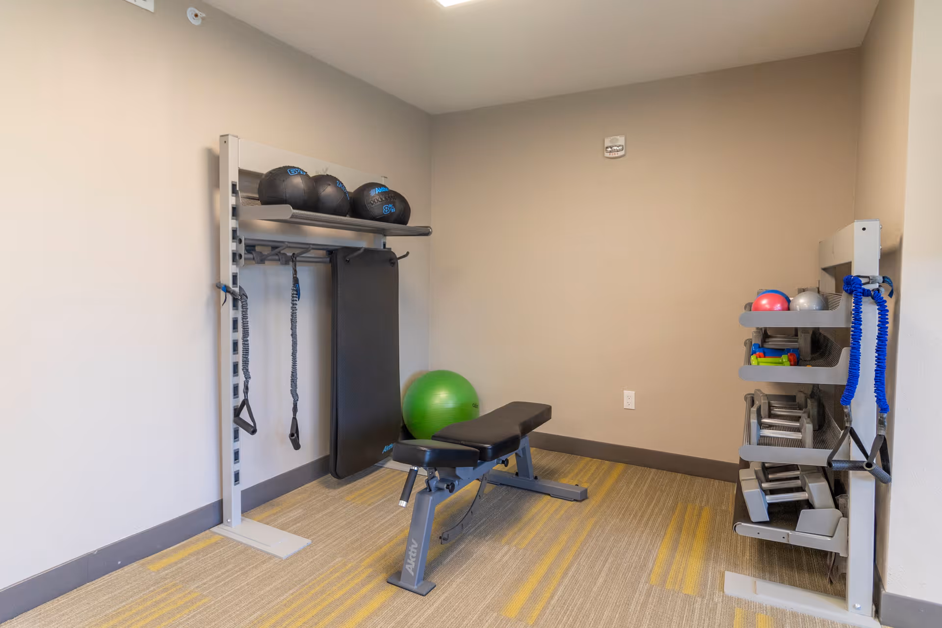 A small fitness room with exercise equipment including a workout bench, medicine balls on a rack, resistance bands hanging on the rack, a green exercise ball in the corner, and dumbbells on shelves against the wall.