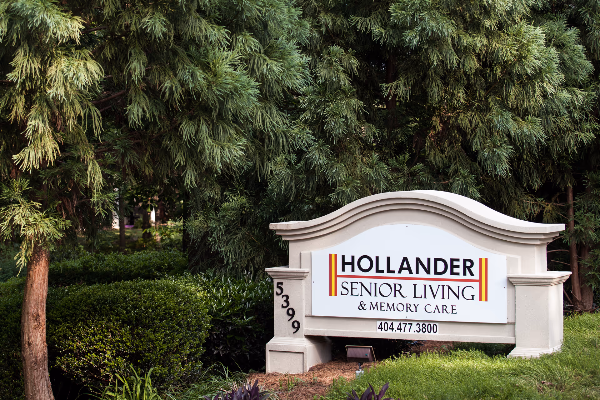 Entrance sign for Hollander Senior Living & Memory Care surrounded by landscaping and trees.