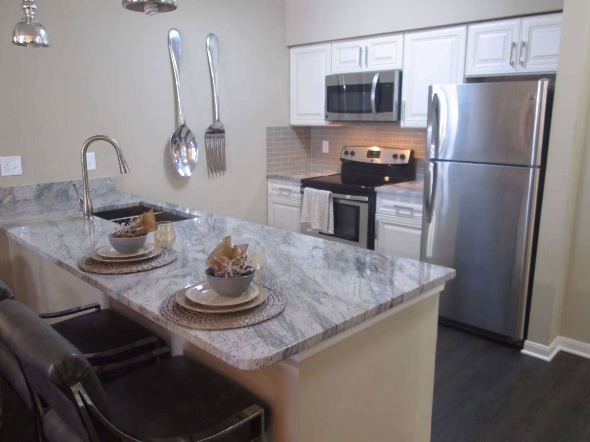 Modern kitchen with a marble island countertop, two bar stools, stainless steel refrigerator and oven, and oversized decorative fork and spoon on the wall.