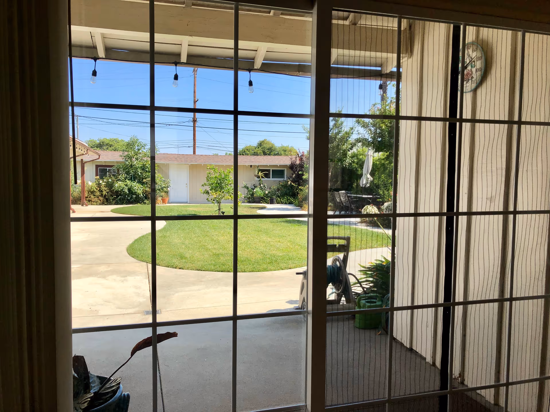View through a glass sliding door with grid patterns, looking out onto a patio area with a circular lawn, some plants, and a building in the background under a clear blue sky.