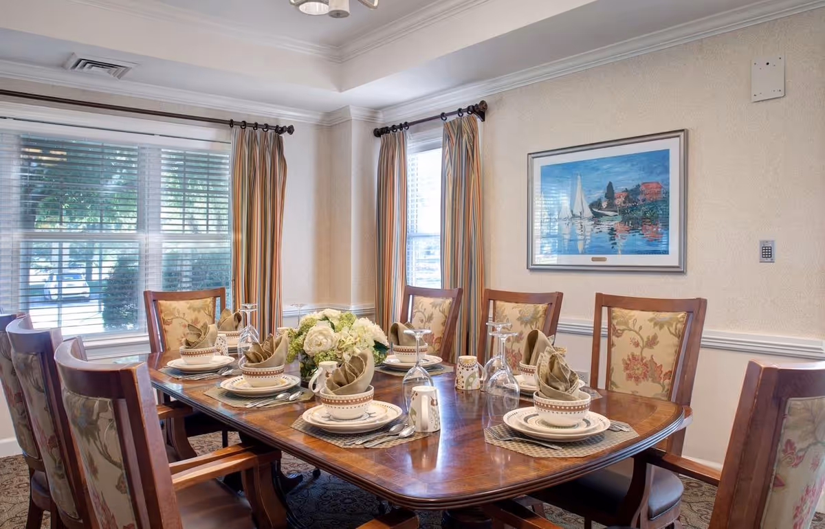 Formal dining room with a set wooden table for six, floral centerpiece, upholstered chairs, large windows with blinds and curtains, and a framed painting on the wall.