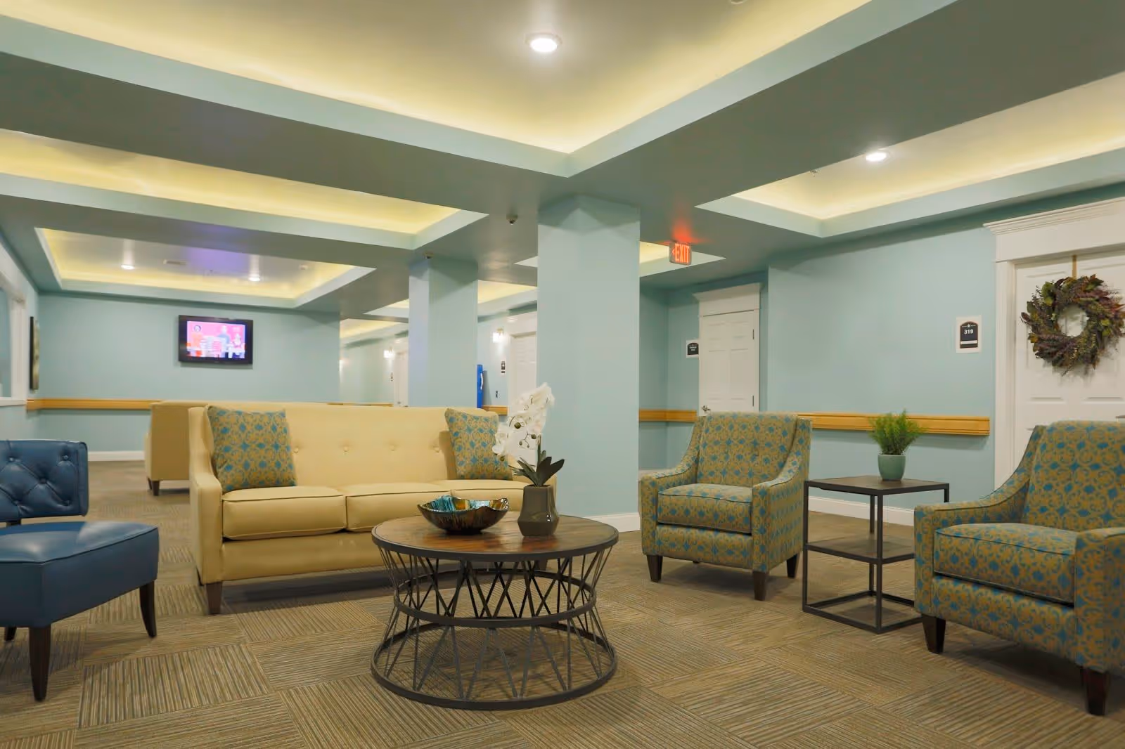 A well-lit common area in a senior living facility with light blue walls and beige carpet. The room features a beige sofa with patterned cushions, two matching patterned armchairs, a blue armchair, a round coffee table with decorative items, and a side table with a small plant. There is a TV mounted on the far wall and a door decorated with a wreath.