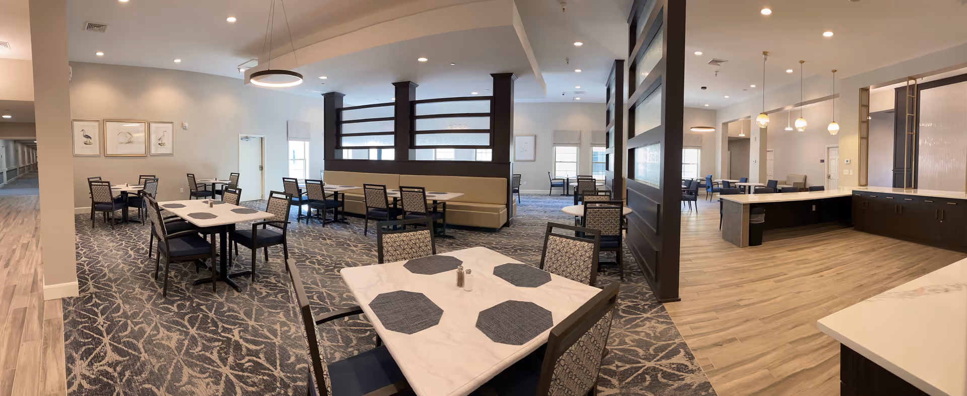 A spacious dining area in a senior living facility featuring multiple tables with chairs and placemats. The room has a patterned carpet, light-colored walls, and modern lighting fixtures. There is a partial divider with frosted glass panels separating the dining area from a kitchen or serving area with wooden flooring and pendant lights.