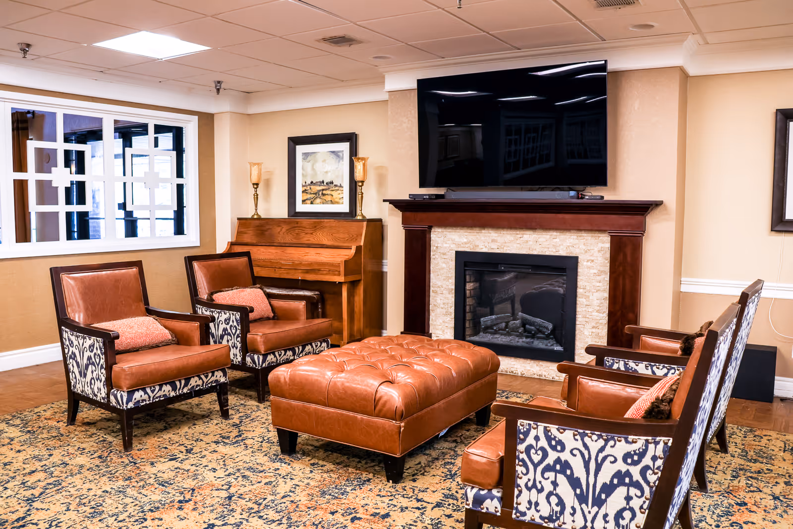 A cozy senior living room with four patterned armchairs and a large tufted leather ottoman arranged around a fireplace with a mounted flat-screen TV above it. There is a wooden piano against the wall with two decorative lamps and a framed picture above it. The room has beige walls and a patterned area rug on the floor.