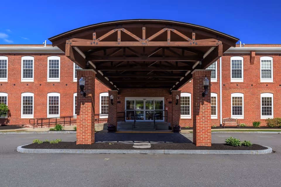 Entrance of a brick building with a covered driveway supported by brick pillars and wooden beams, with multiple windows and a clear blue sky in the background.