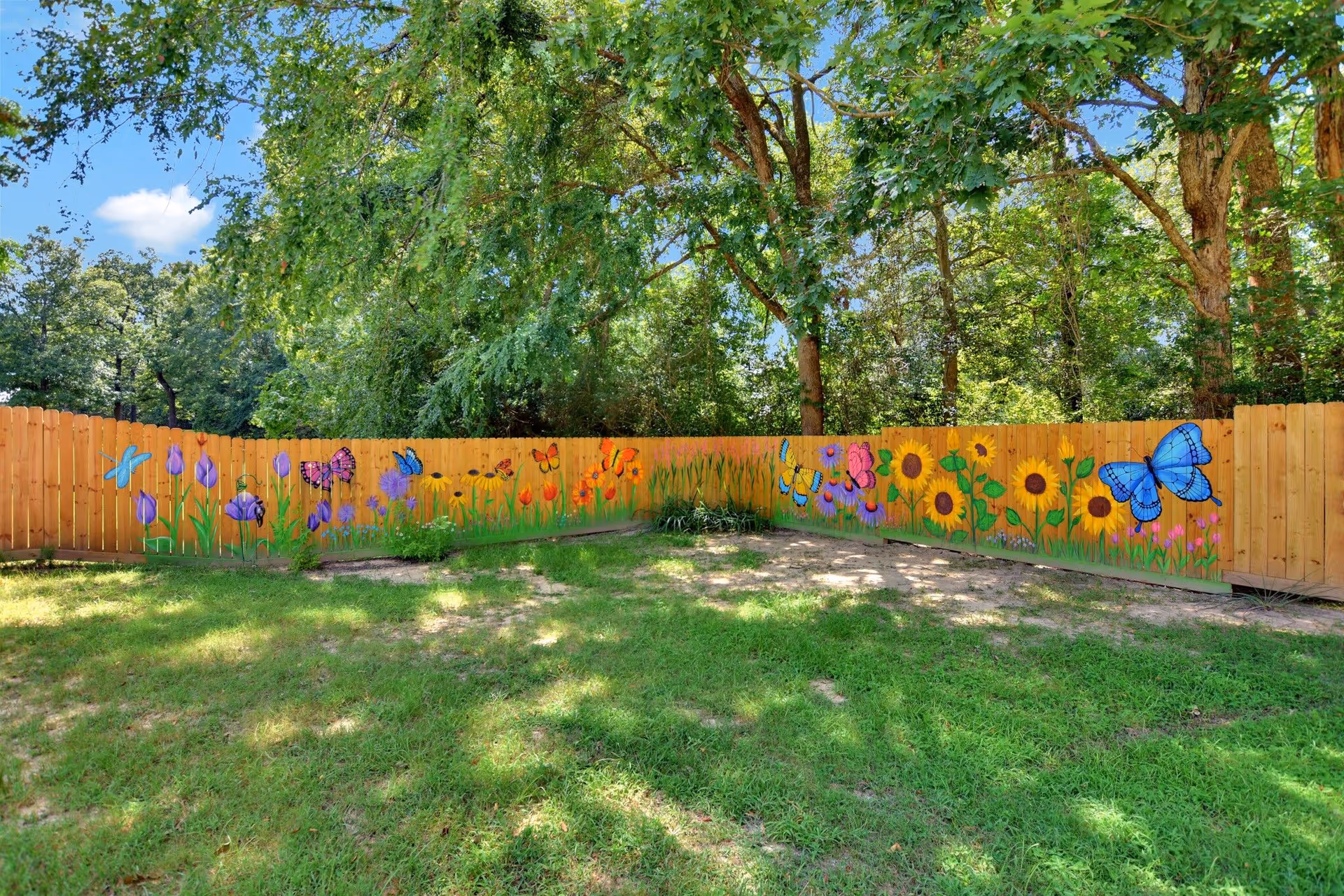 A wooden fence painted with colorful flowers and butterflies in a grassy outdoor area with trees in the background under a blue sky with some clouds.