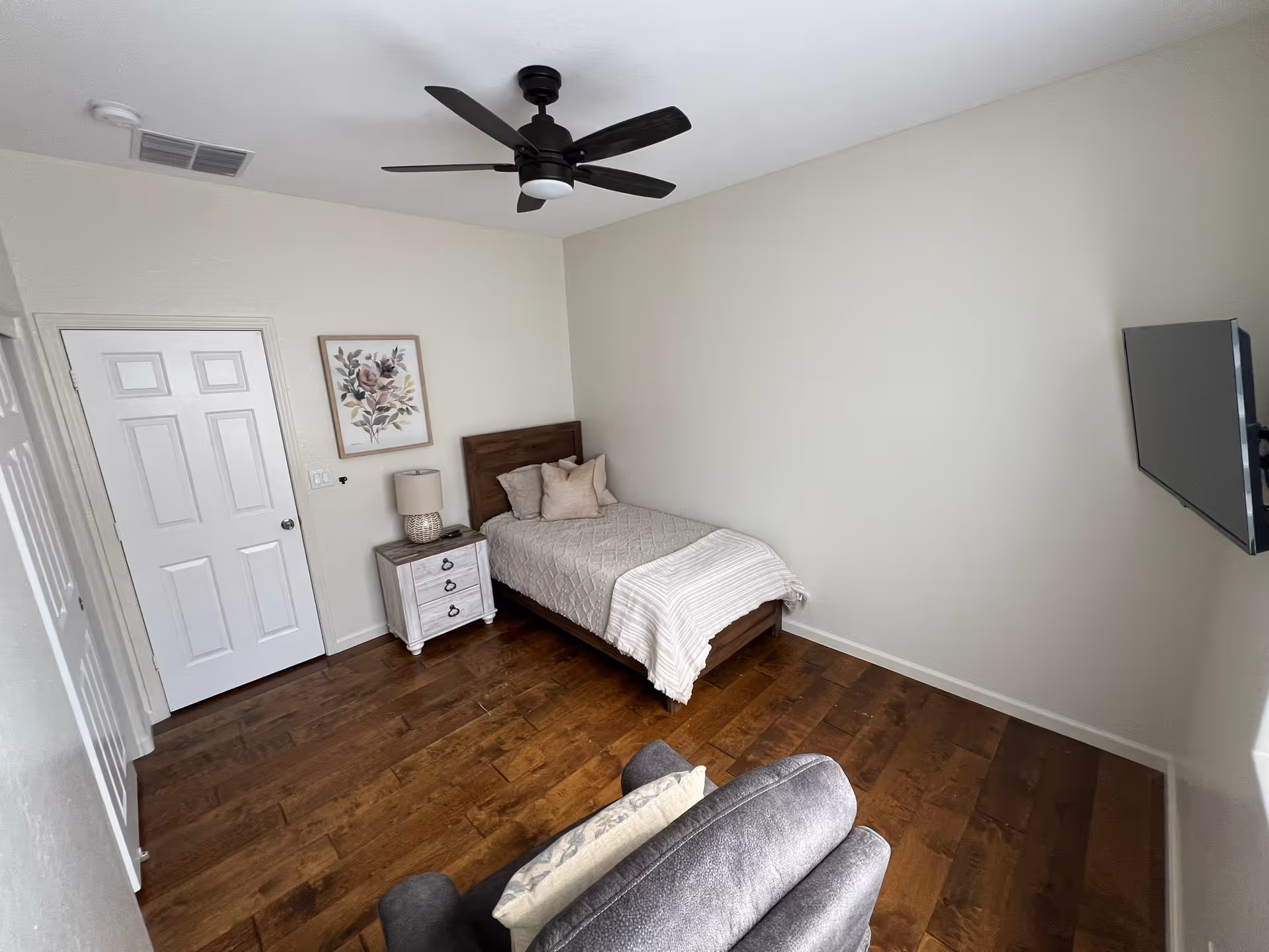 Small, minimally furnished bedroom with a single bed, nightstand and lamp, ceiling fan, wall-mounted TV, and an armchair on hardwood floors.