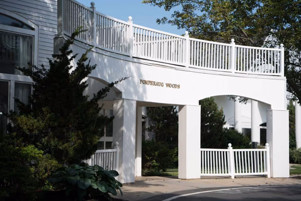 Entrance archway of Pomperaug Woods facility with white railings and columns, surrounded by greenery and trees under a clear blue sky.