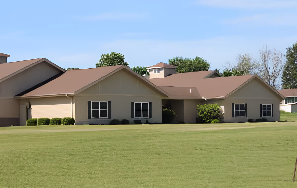 Exterior view of a single-story building with beige walls and brown roofs, surrounded by a well-maintained green lawn and some bushes, under a clear blue sky.