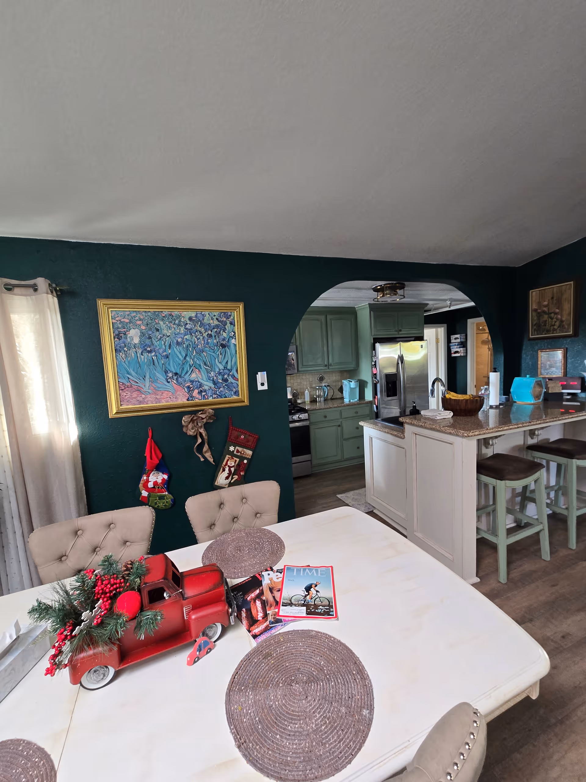 A dining area with a white table and beige cushioned chairs. On the table, there is a red decorative truck with greenery and berries, along with magazines. The wall behind the table is dark green with a framed painting and two Christmas stockings hanging. Through an arched opening, a kitchen with green cabinets, a stainless steel refrigerator, and a granite countertop with bar stools is visible.