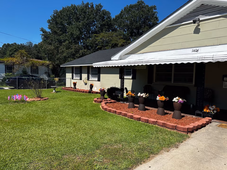 Front exterior of a single-story house with a covered porch, multiple flower pots in a mulched bed and a well-kept lawn under a clear blue sky.