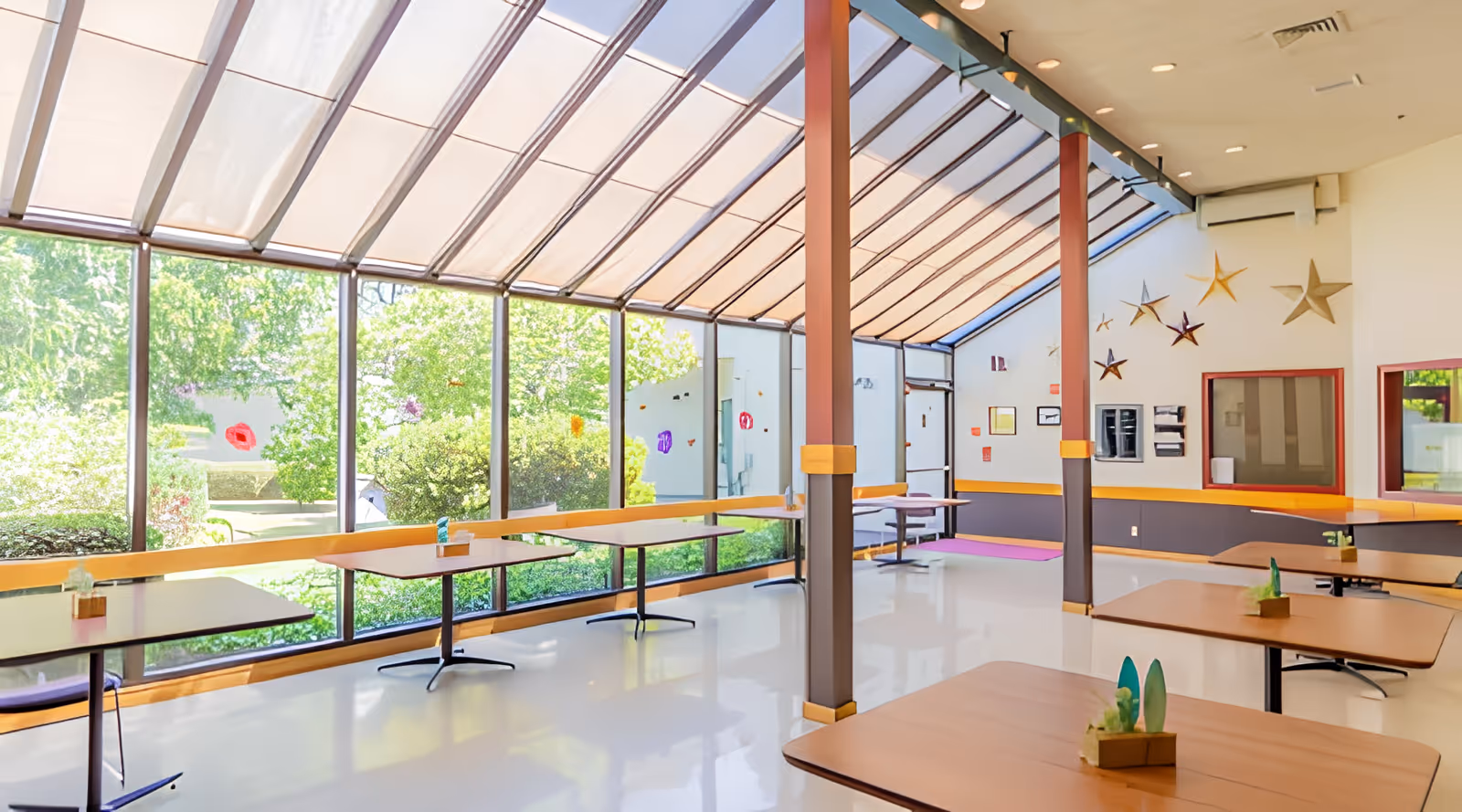 Bright dining area with large floor-to-ceiling windows and a slanted glass ceiling letting in natural light. Several tables with small decorative plants are spaced throughout the room. The walls are decorated with star-shaped ornaments, and there is a view of green bushes and trees outside.