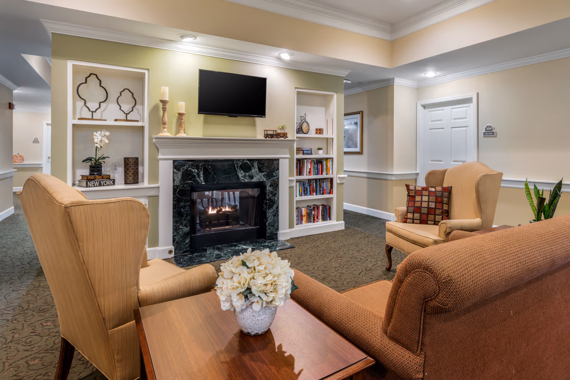A cozy living room area in a senior living facility featuring a fireplace with a black marble surround and a flat-screen TV mounted above it. The room has two beige wingback chairs, a brown patterned sofa, and a wooden side table with a white flower arrangement. Built-in shelves flank the fireplace, holding decorative items and books. The walls are painted light beige with white trim, and the carpet has a subtle pattern. A door labeled '309' is visible in the background.