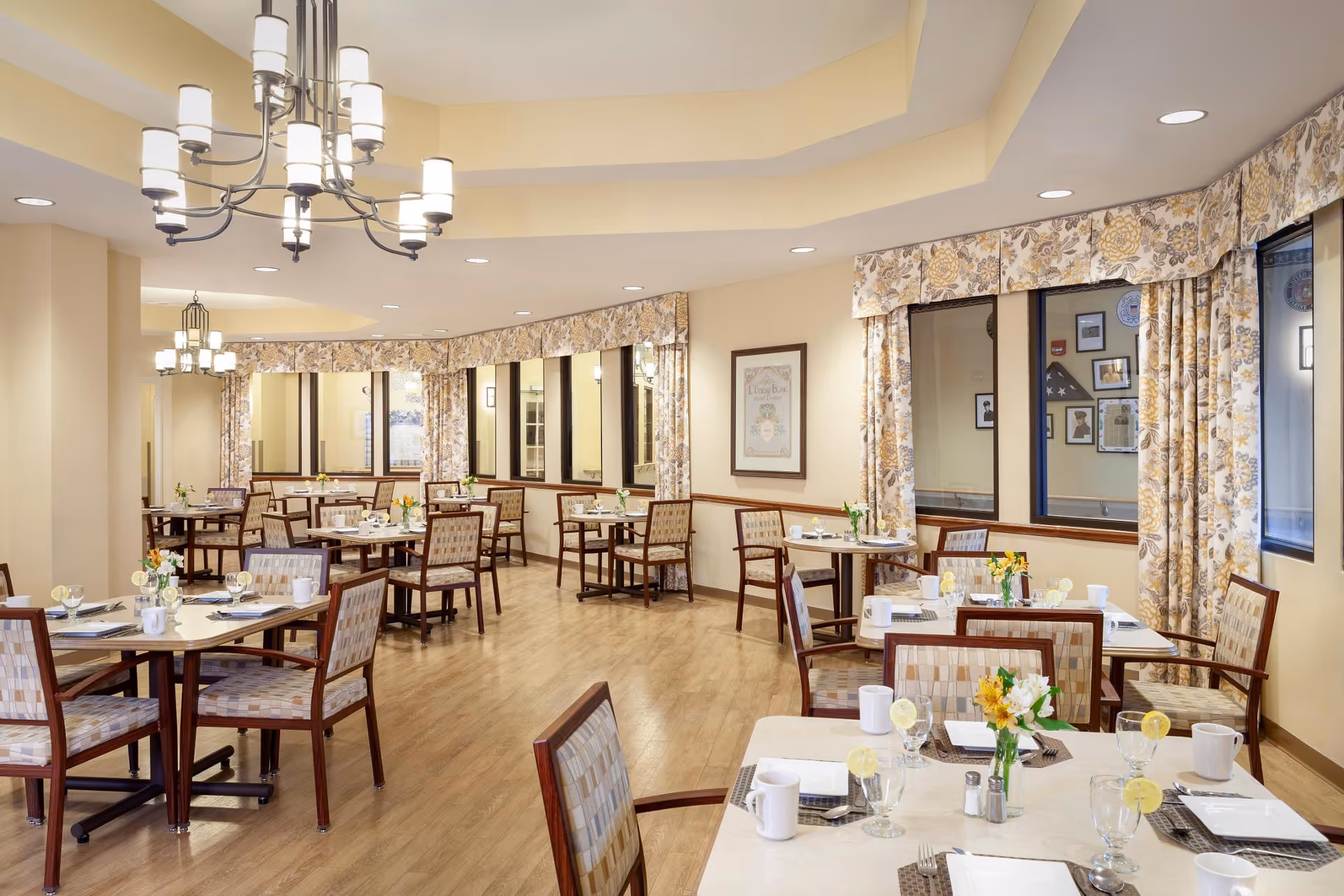Bright dining room with multiple set tables and chairs, chandeliers, wood flooring, and floral curtains.
