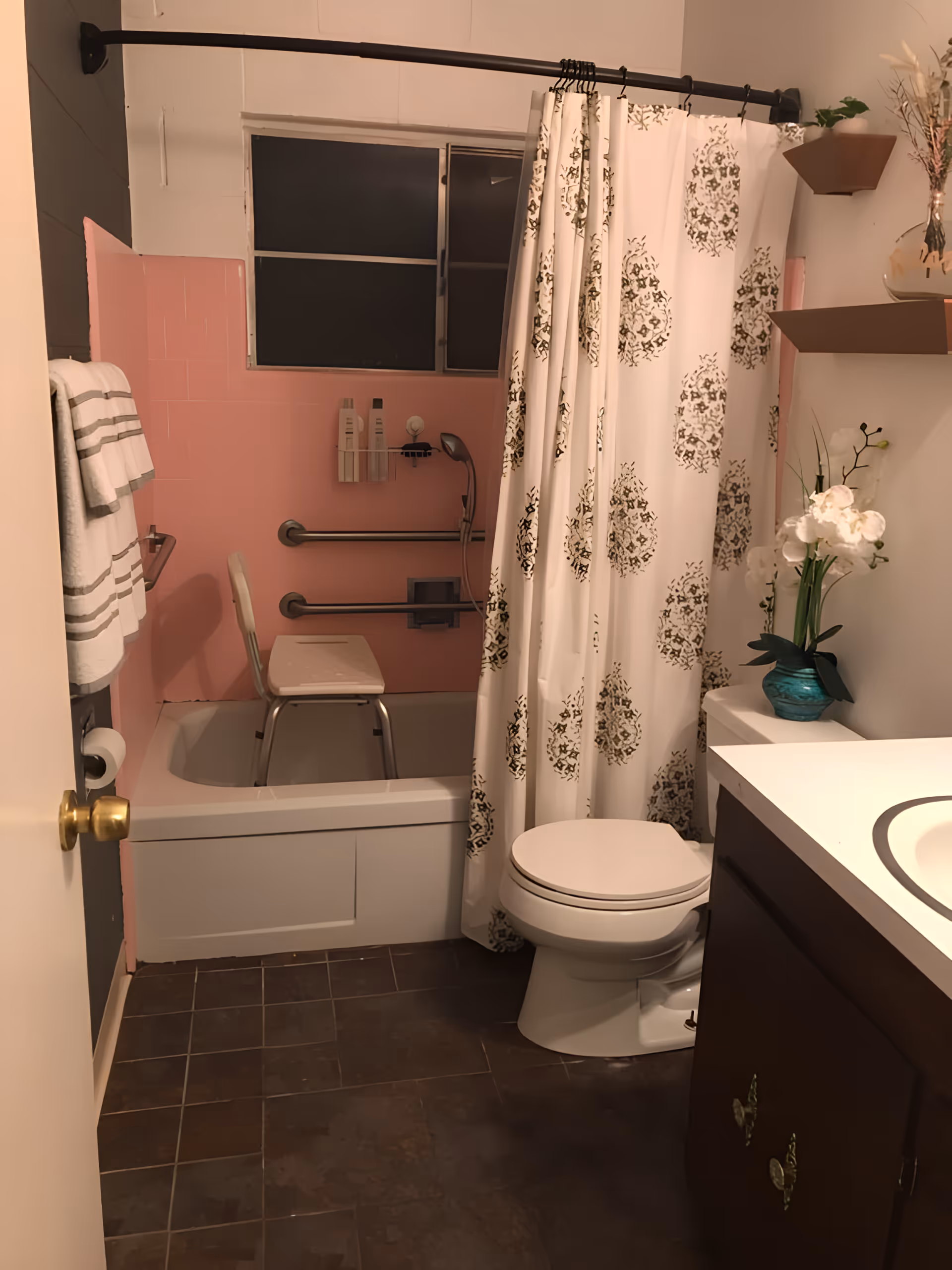 Small bathroom with a tub and shower chair, patterned shower curtain, pink tile wall, toilet, sink vanity, and shelves with a potted orchid.
