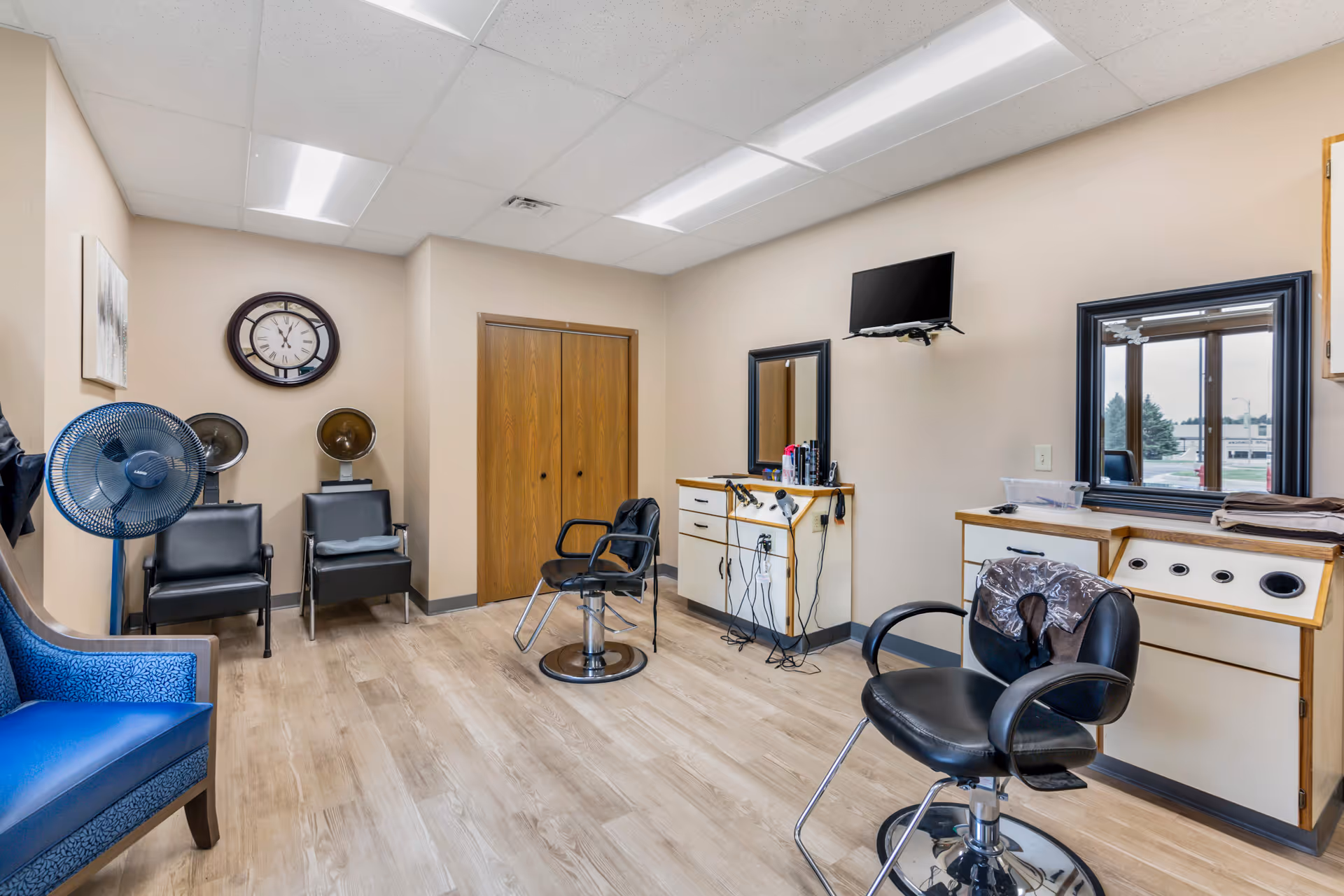 Interior salon/hair-styling room with chairs, mirrors, hair dryers, and a wall-mounted TV.