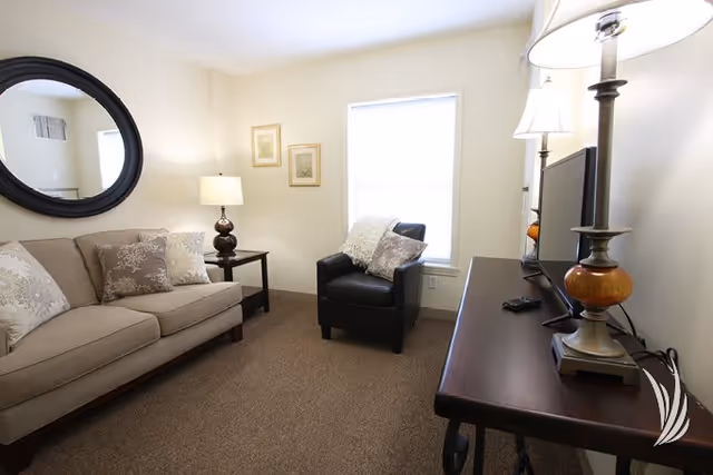 A cozy living room with a beige sofa adorned with patterned pillows, a black armchair with a throw blanket, a dark wooden side table with a lamp, a round black-framed mirror on the wall, two framed pictures, a window letting in natural light, and a dark wooden console table holding a TV and a lamp.