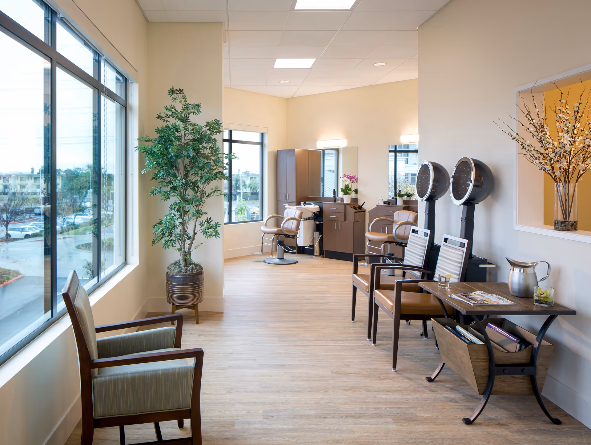 Bright and clean salon area with large windows, wooden flooring, two hair drying chairs with hooded dryers, salon chairs, a potted plant, and a small table with magazines and a pitcher of water.