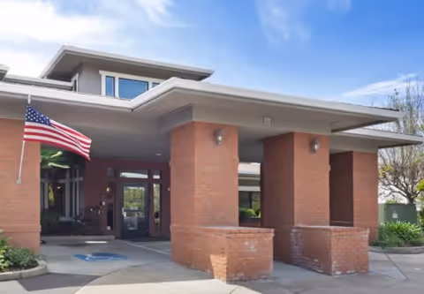 Covered brick entrance with square columns, a mounted American flag, and glass double doors.