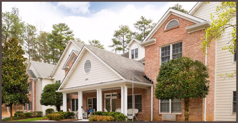 Exterior view of a senior living facility building with brick and siding walls, a covered porch with white columns, and neatly trimmed bushes and trees in front.