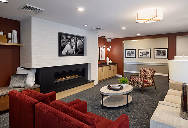 A cozy senior living facility common area with a modern fireplace set in a white tiled wall. The room features a red sofa, a patterned armchair, a beige sofa, a round coffee table with a small plant and books, and framed black-and-white photos on the walls. The lighting includes ceiling lights and wall sconces, creating a warm and inviting atmosphere.