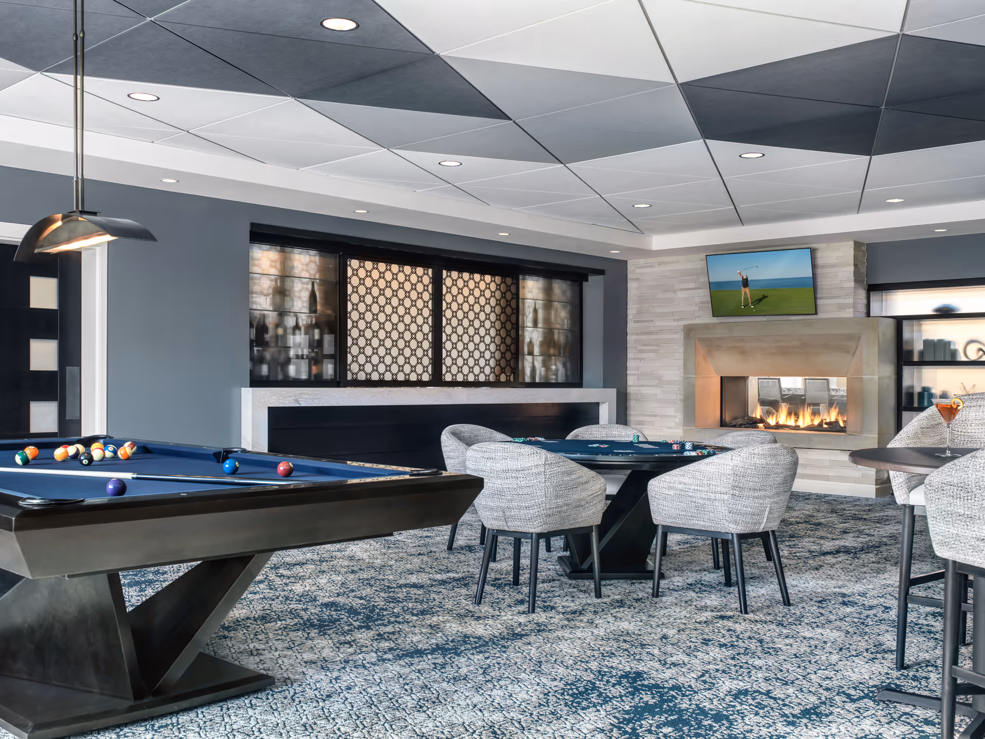 A modern game room with a pool table, several round tables with gray upholstered chairs, a lit fireplace, and a wall-mounted TV showing a golfer. The room features a patterned carpet, geometric ceiling tiles, and a decorative window with a lattice design.