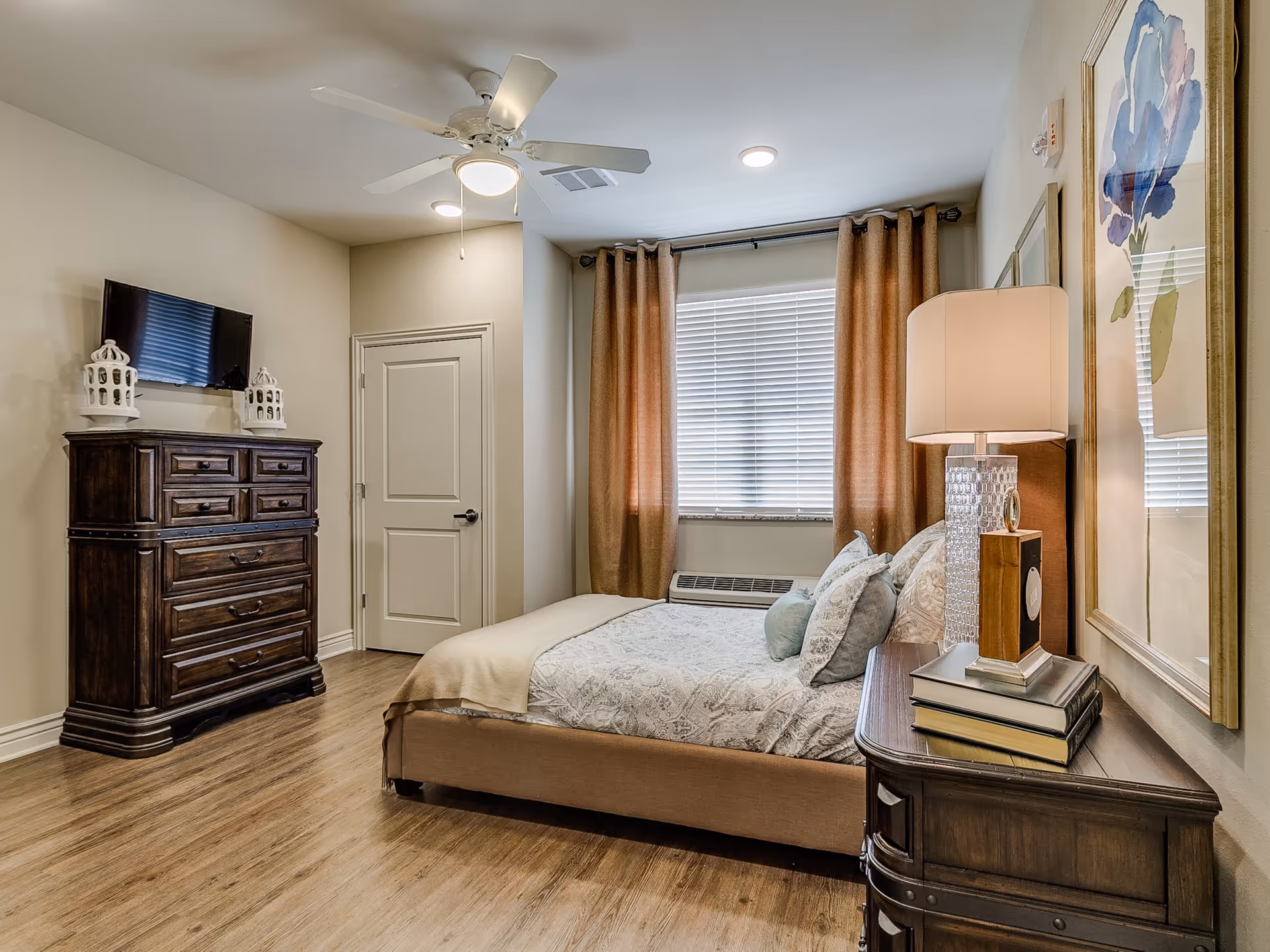 A cozy bedroom with a bed featuring patterned bedding and multiple pillows. There is a wooden nightstand with a lamp, books, and a decorative clock. A large window with closed blinds and brown curtains is behind the bed. Across from the bed is a wooden dresser with decorative lanterns and a wall-mounted flat screen TV. The room has wood flooring, a ceiling fan with light, and neutral-colored walls.