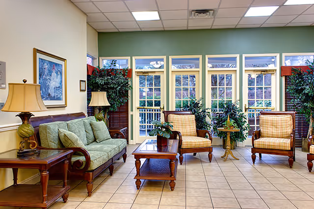 Sunlit communal lounge with a green sofa, plaid armchairs, wooden coffee tables, plants, and large windows/doors to an outdoor area.