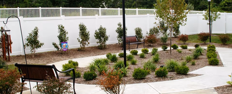 Curved paved courtyard with benches, planted shrubs and small trees enclosed by a white privacy fence.