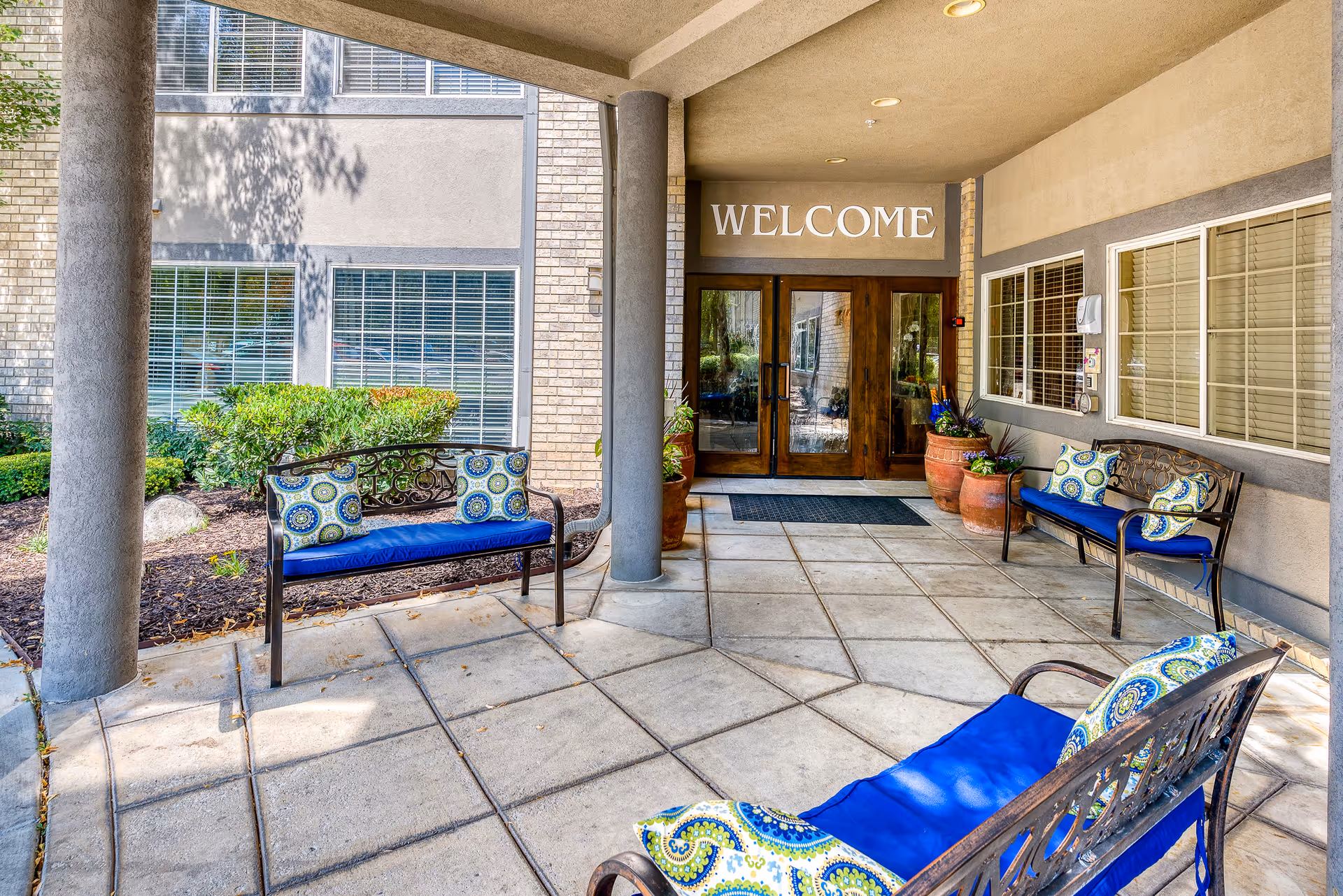 Covered building entrance with double glass doors beneath a 'WELCOME' sign and decorative benches with blue cushions.