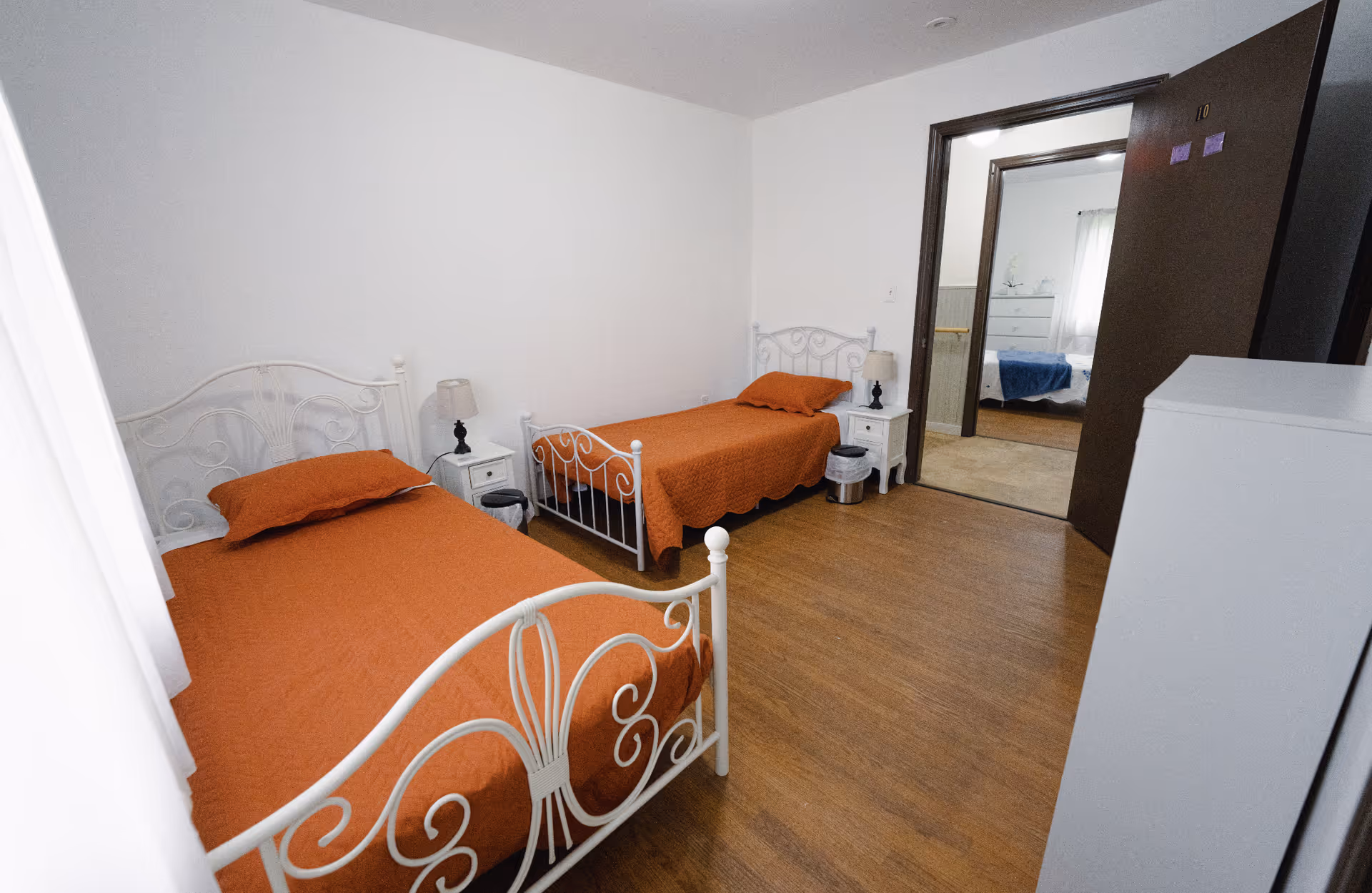A simple bedroom with two single beds featuring white metal frames and orange bedspreads. Each bed has a matching orange pillow. Between the beds are two small white nightstands with lamps. The room has wooden flooring and white walls. An open door reveals another room with a bed covered in a blue blanket.