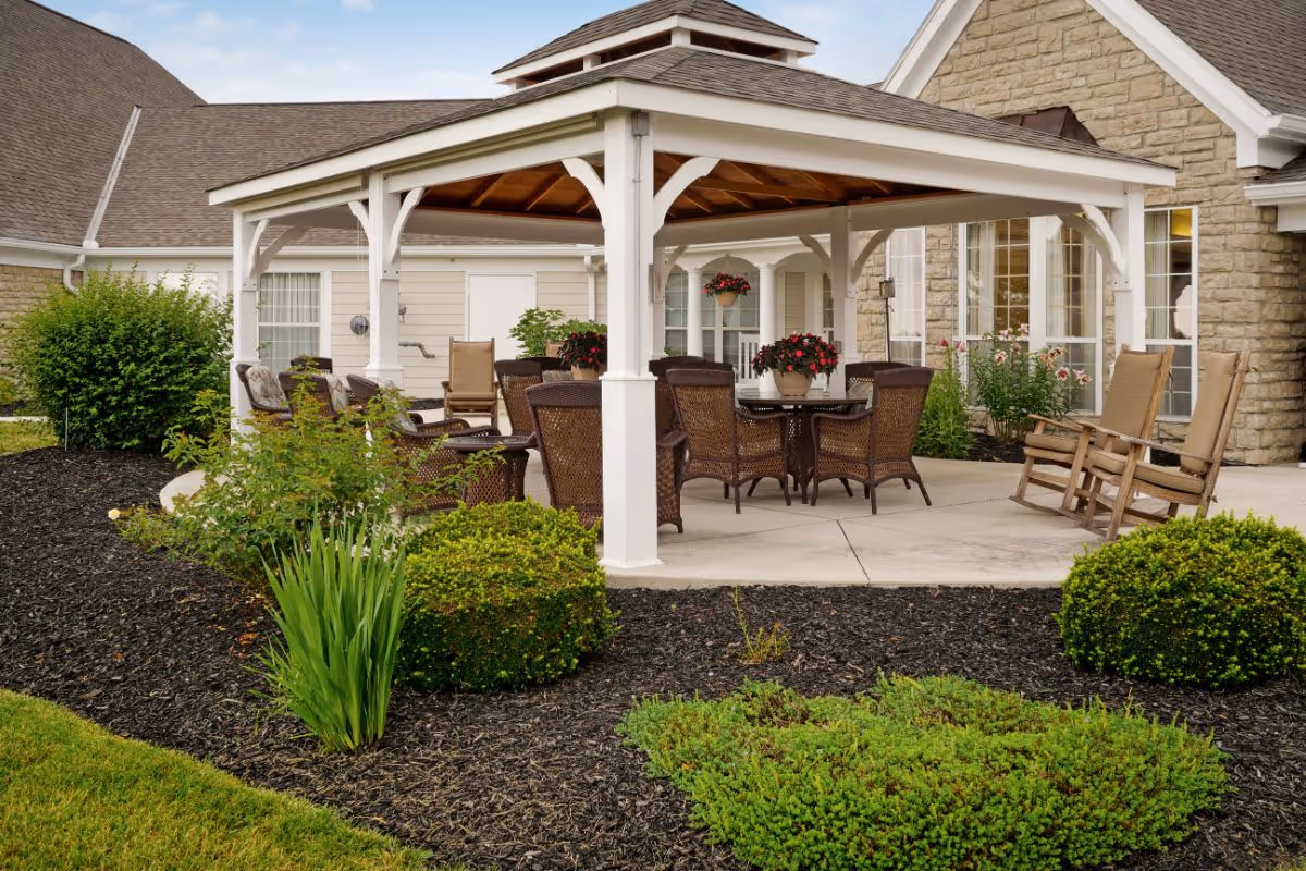 Outdoor patio area at Sunrise of Findlay featuring a white wooden gazebo with a brown roof. Under the gazebo are several brown wicker chairs around a round table with potted flowers. Two wooden rocking chairs are placed on the concrete patio outside the gazebo. The surrounding area has well-maintained landscaping with green bushes, plants, and mulch beds. The building exterior is visible in the background with beige siding and stone accents.