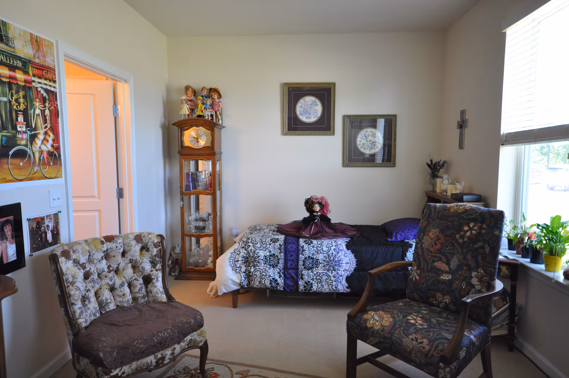 A furnished bedroom with a single bed topped by a decorative doll, two upholstered chairs, a glass-front clock cabinet, framed wall art, and a window with potted plants.