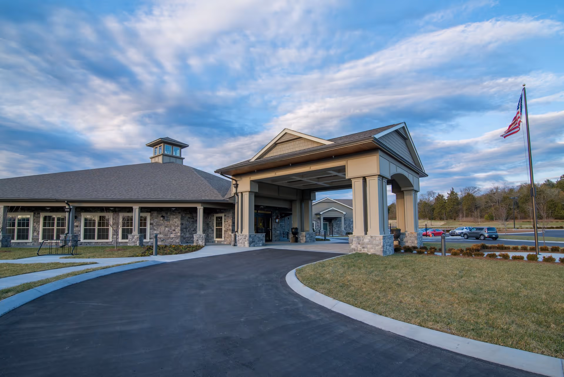 Exterior view of Morning Pointe of Franklin facility showing a large covered entrance with stone and beige siding, a curved driveway, a flagpole with an American flag, and a parking area with several cars under a partly cloudy sky.