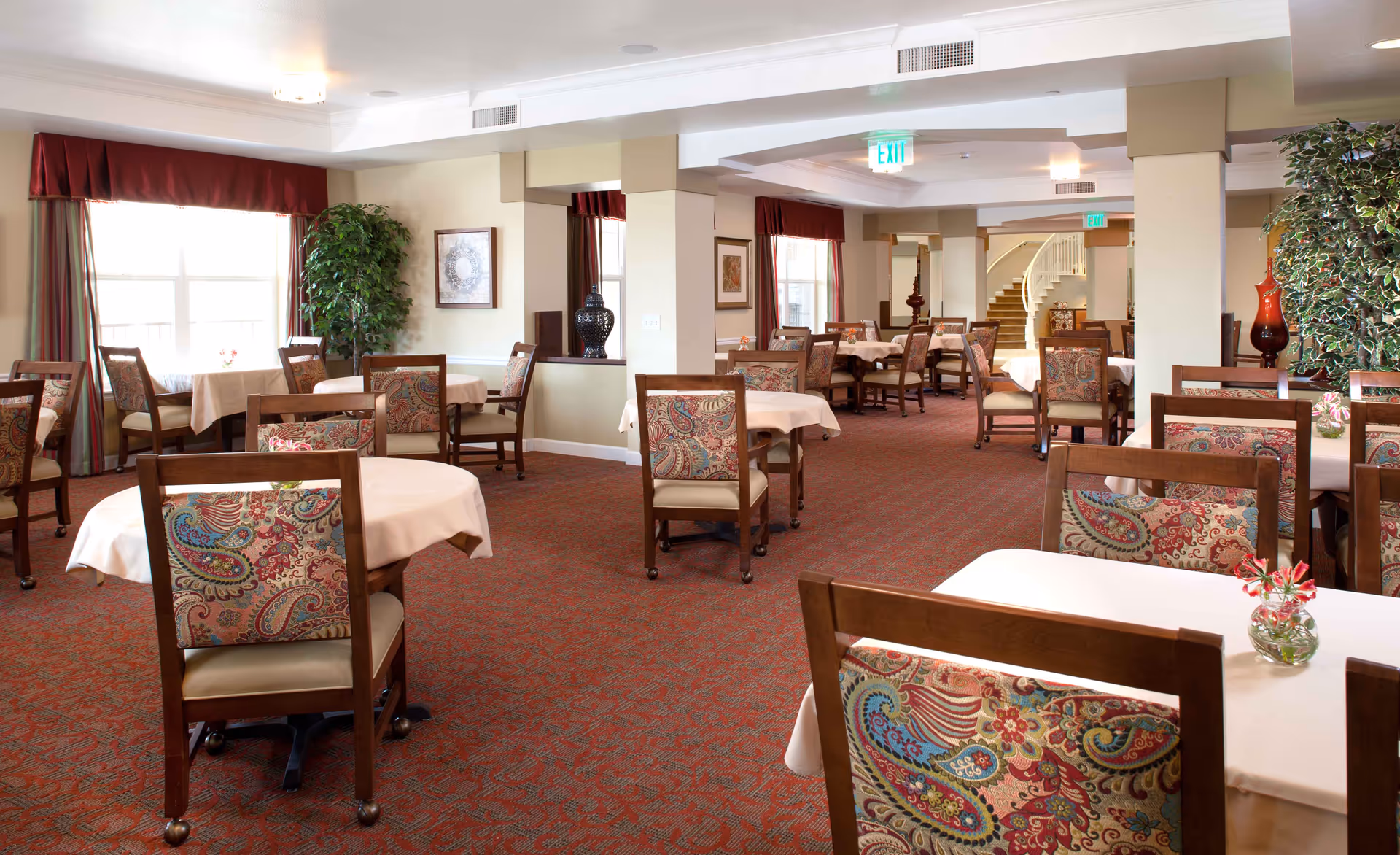 A spacious dining room with multiple tables covered in white tablecloths and wooden chairs with colorful patterned upholstery. The room has large windows with red curtains, potted plants, framed artwork on the walls, and a carpeted floor with a red and orange pattern. There is a staircase visible in the background.