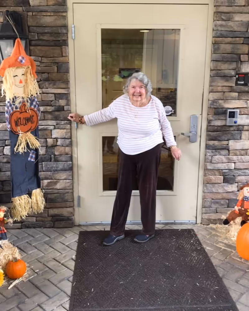 An elderly woman with gray hair, wearing a white and pink striped long-sleeve shirt and dark pants, stands smiling in front of a beige door with glass panels. The door is set in a stone wall. To the left of the door is a scarecrow decoration holding a pumpkin-shaped sign that says 'Welcome'. On the ground near the door are small pumpkins and hay decorations.
