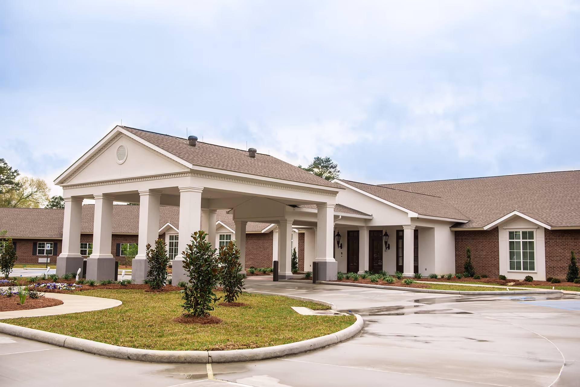 Front entrance of a single-story senior care building with a covered porte-cochère, columns, and landscaped driveway.