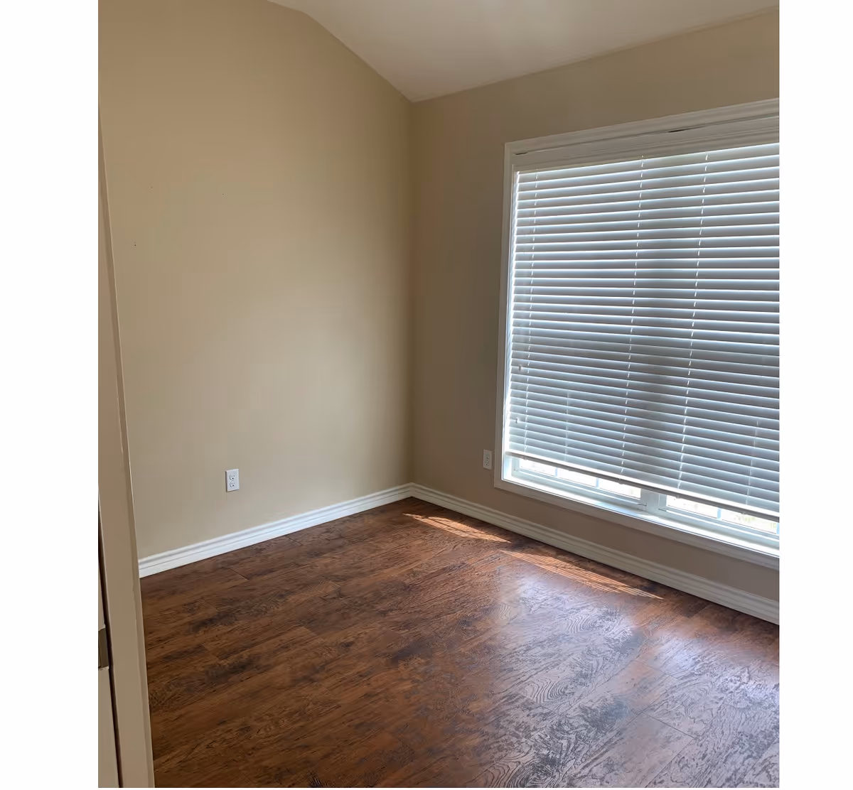Empty small room with hardwood floors, beige walls, and a large window with closed blinds.