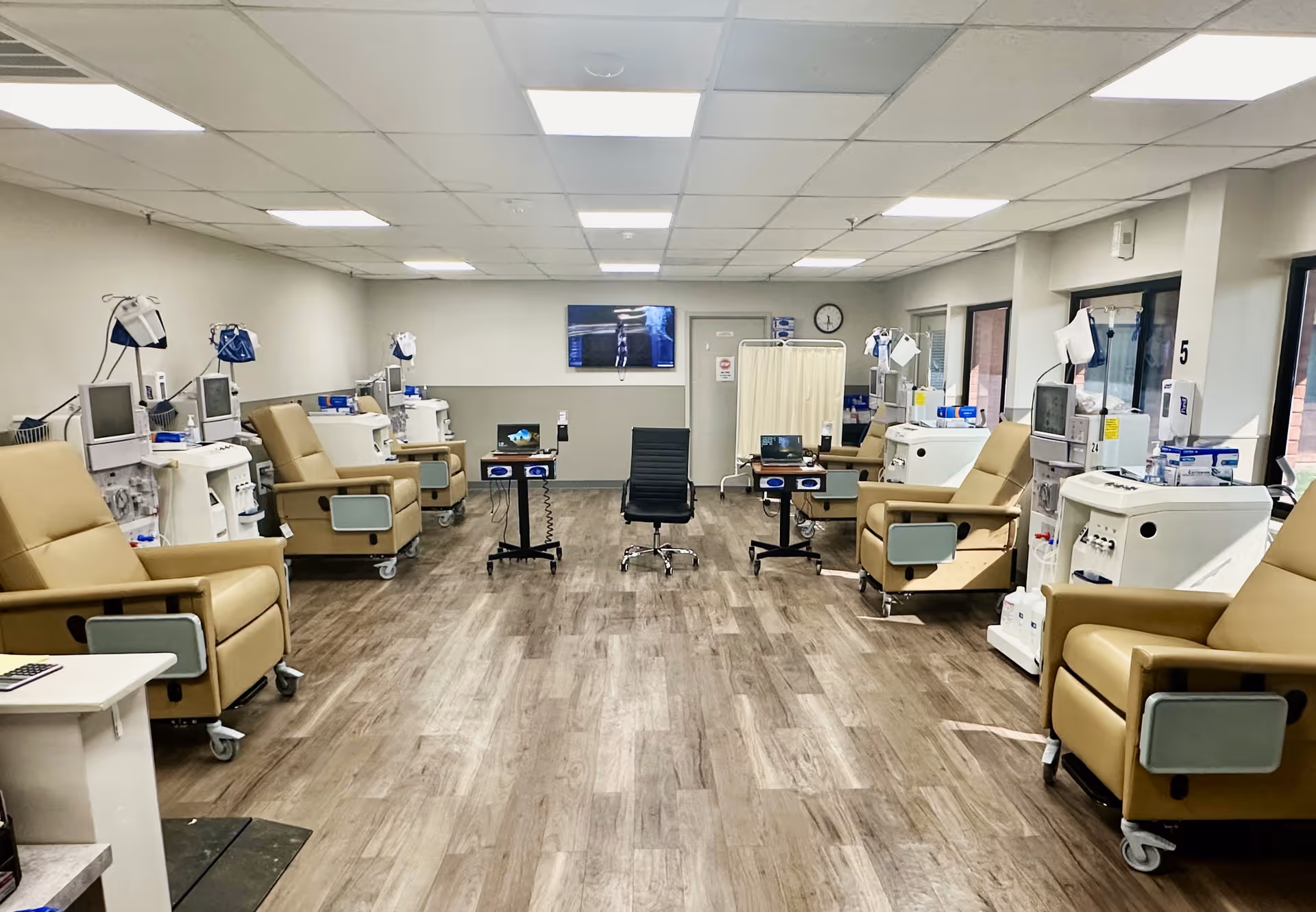 A medical treatment room with multiple beige recliner chairs arranged along both sides of the room. Each chair is paired with medical equipment, including dialysis machines and IV stands. The room has wood-patterned flooring, white walls, and a drop ceiling with fluorescent lighting. A black office chair and two small tables with medical devices are positioned in the center. A TV is mounted on the far wall above a door, and windows line the right side of the room.