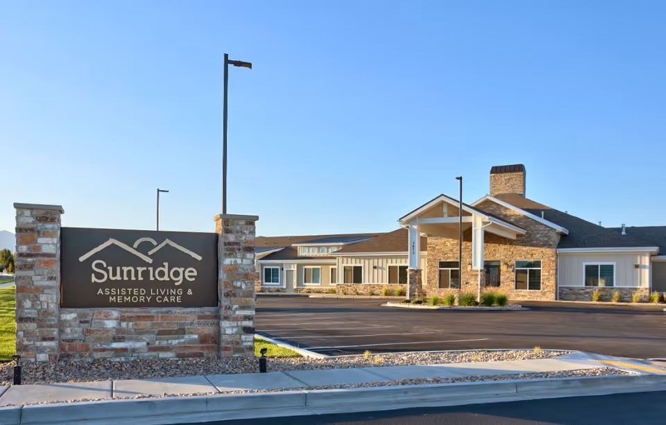 Exterior view of Sunridge Assisted Living & Memory Care facility in Roy, showing a single-story building with stone and siding facade, a covered entrance, and a parking lot in front under a clear blue sky.