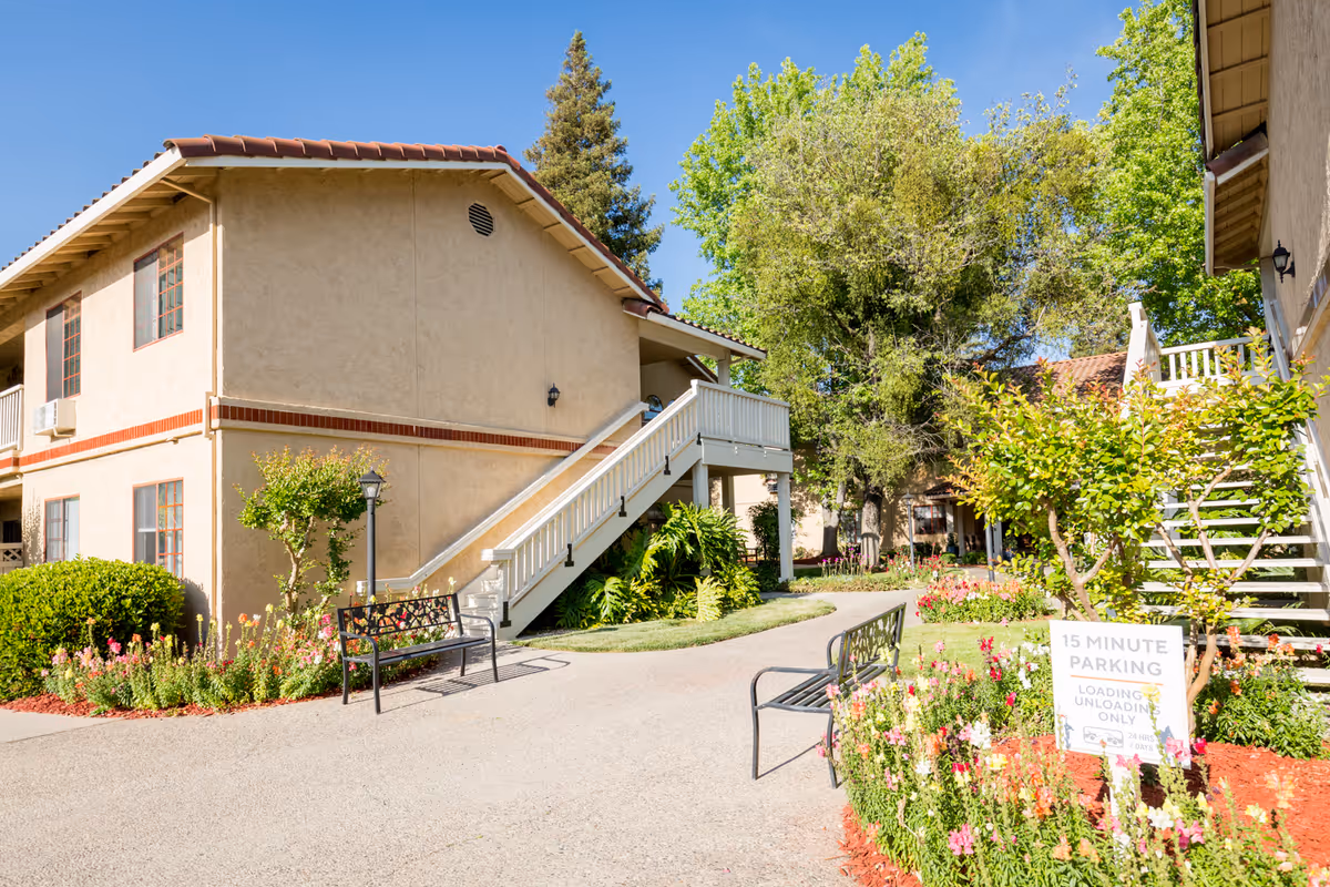 Outdoor view of a senior living facility courtyard with beige two-story buildings featuring red-tiled roofs. There are stairs leading to the second floor, benches along the walkway, and colorful flower beds with green shrubs and trees under a clear blue sky.