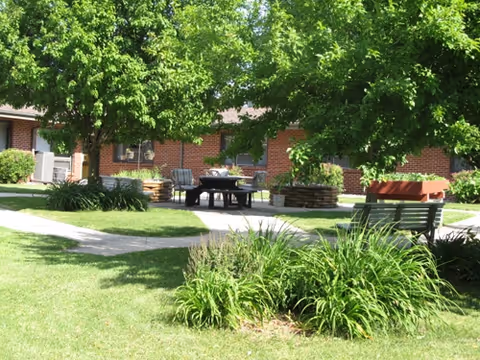Outdoor seating area with benches and a table surrounded by green grass, plants, and trees in front of a brick building under bright sunlight.