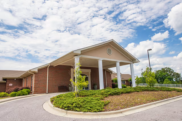 Brick senior living facility entrance with a portico supported by white columns, circular driveway, and a landscaped island under a partly cloudy sky.