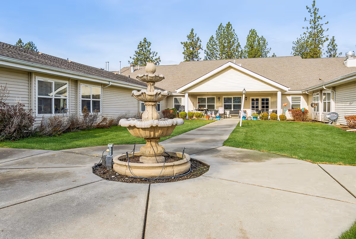 A circular courtyard with a multi-tiered stone fountain in front of a single-story senior living building with a covered entrance and green lawn.